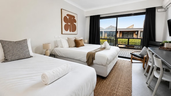 Une chambre d’hôtel avec deux lits, un bureau et une vue sur un immeuble, visible à travers de grandes fenêtres.