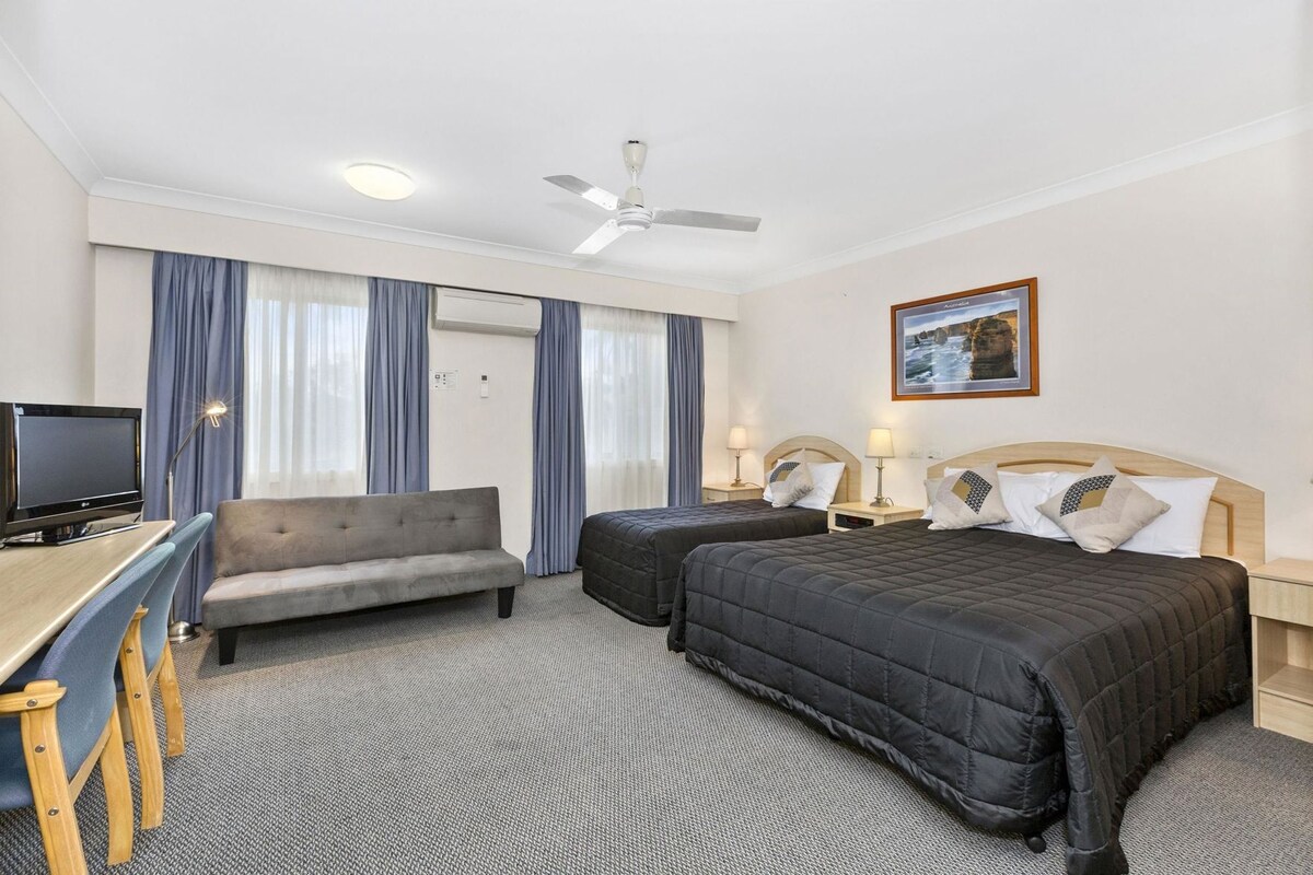 A hotel room with a bed, two bedside tables with lamps, a desk with a computer, a sofa, and a ceiling fan.
