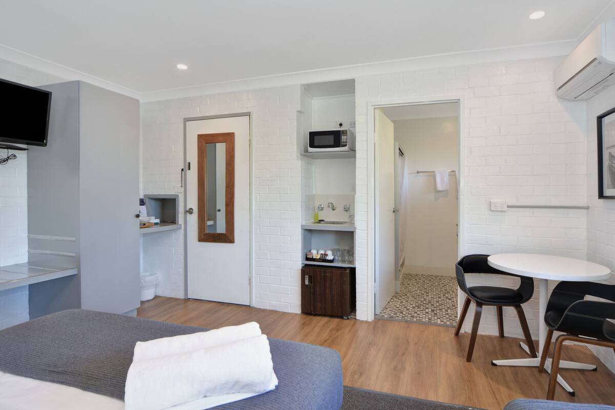 A hotel room with a bed, a TV, a microwave, a small table with chairs, and a bathroom visible through an open door.