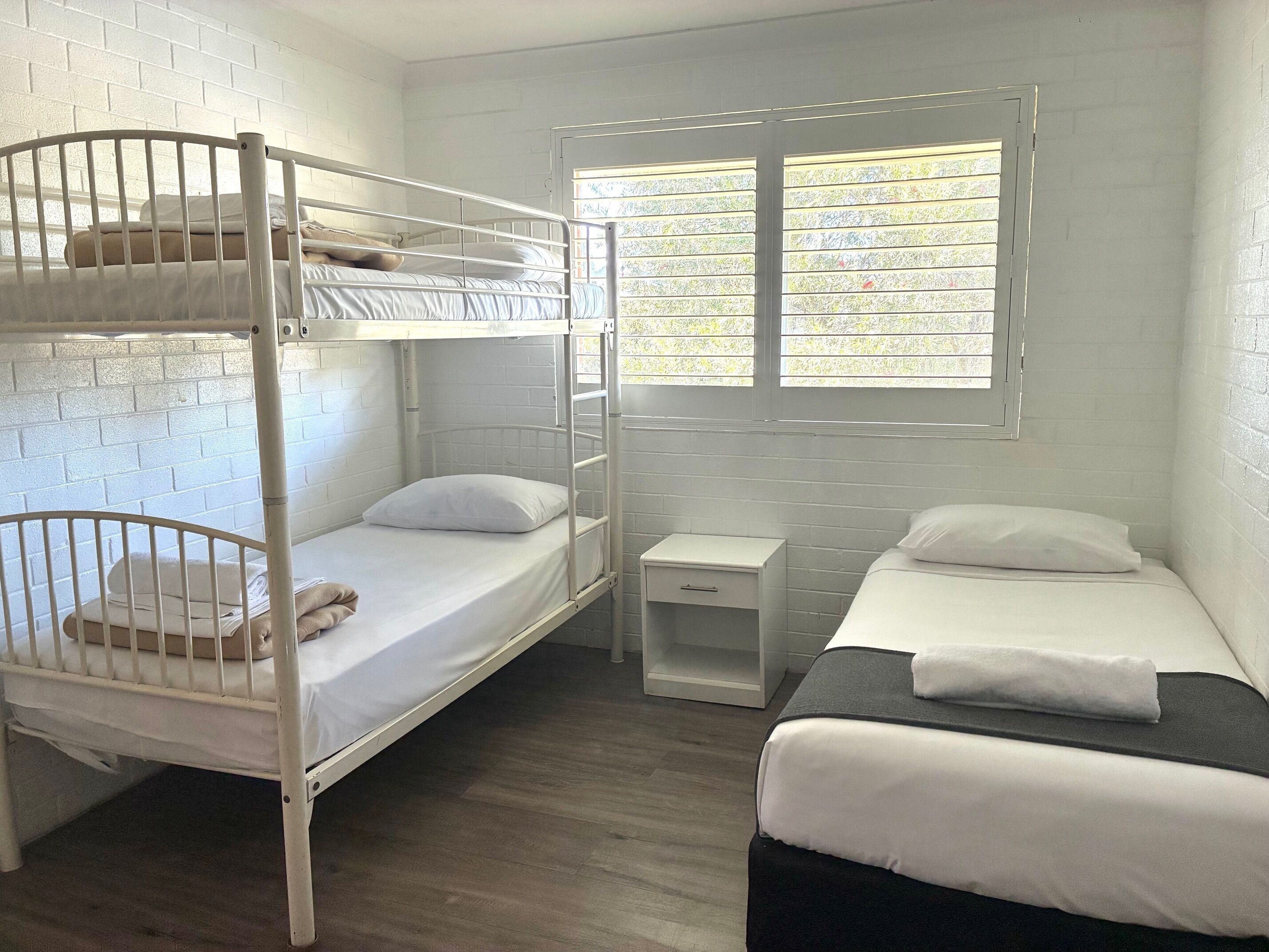 A dormitory room with bunk beds, a small bedside table, and a window with blinds.
