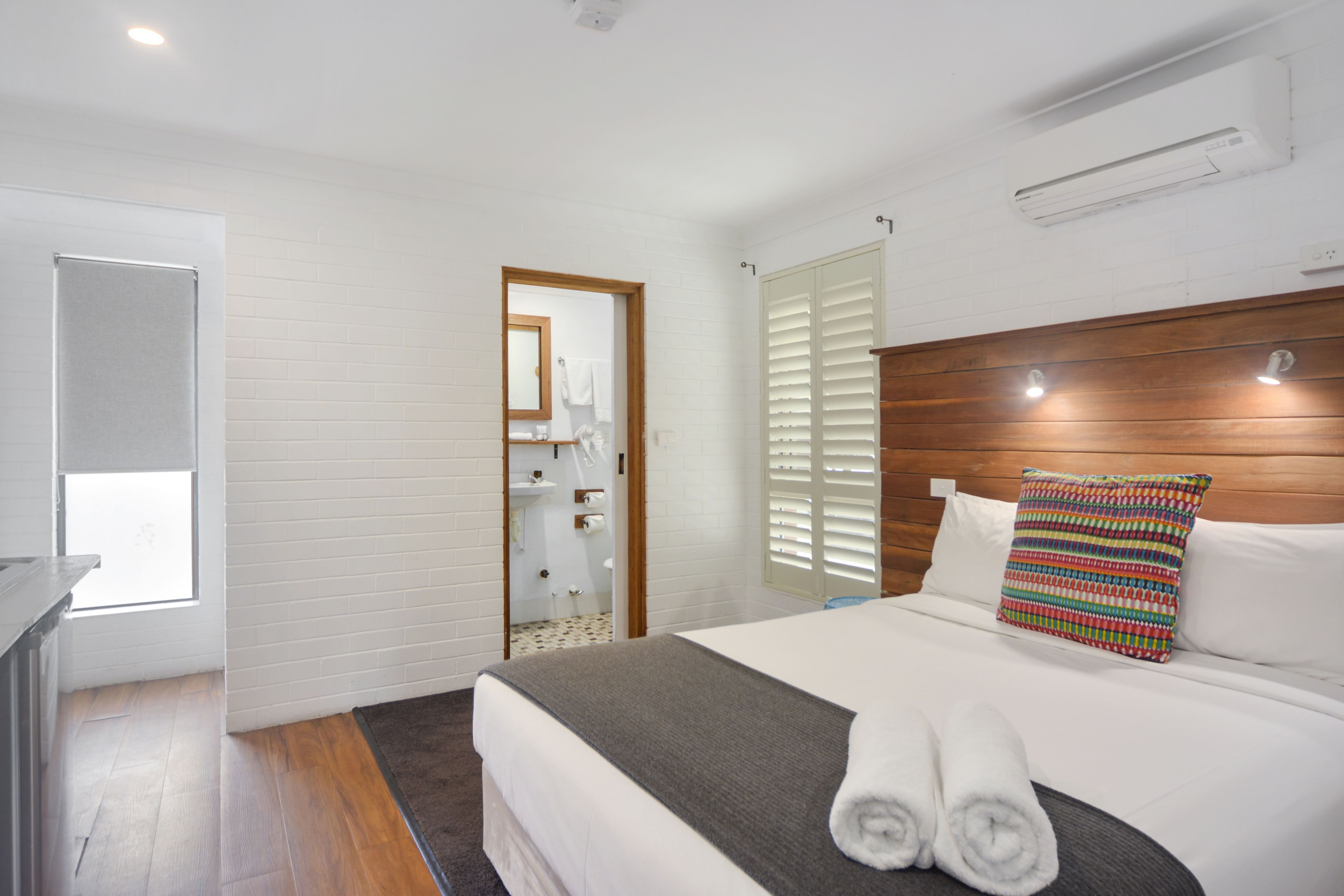 A hotel room with a wooden headboard, a bed with white linens, a colorful pillow, and a view of the bathroom through an open door.