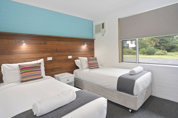 A hotel room with two beds, a wooden headboard, a window with a view of greenery, and a small bedside table with a lamp. - Pleasant Way River Lodge (Nowra)