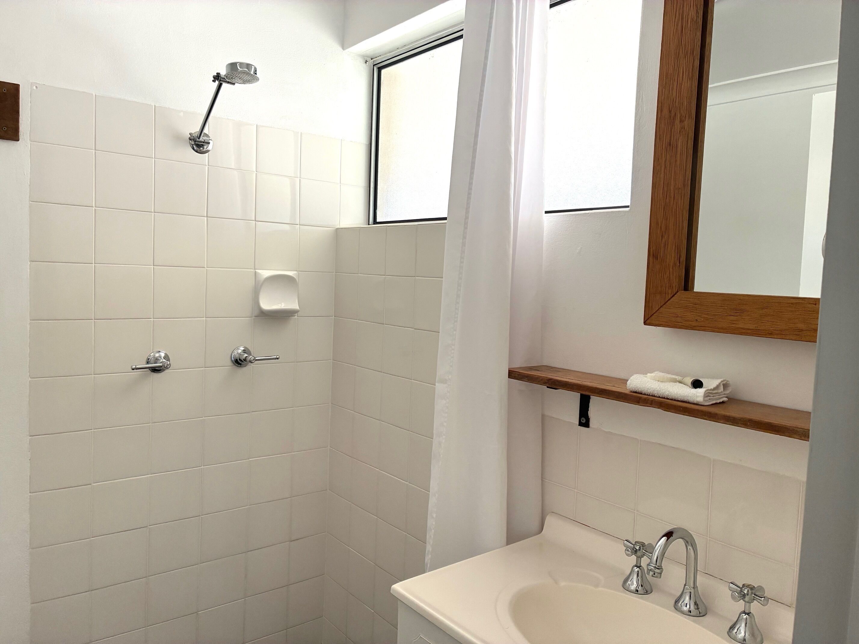 A bathroom with white tiles, a shower, a sink with two faucets, a soap dispenser, and a wooden shelf with a towel.