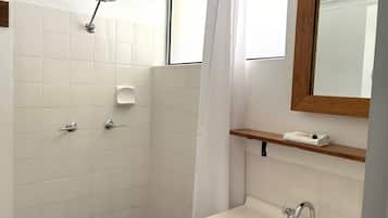 Un baño con azulejos blancos, ducha, lavabo con dos grifos, dispensador de jabón y estante de madera con una toalla.