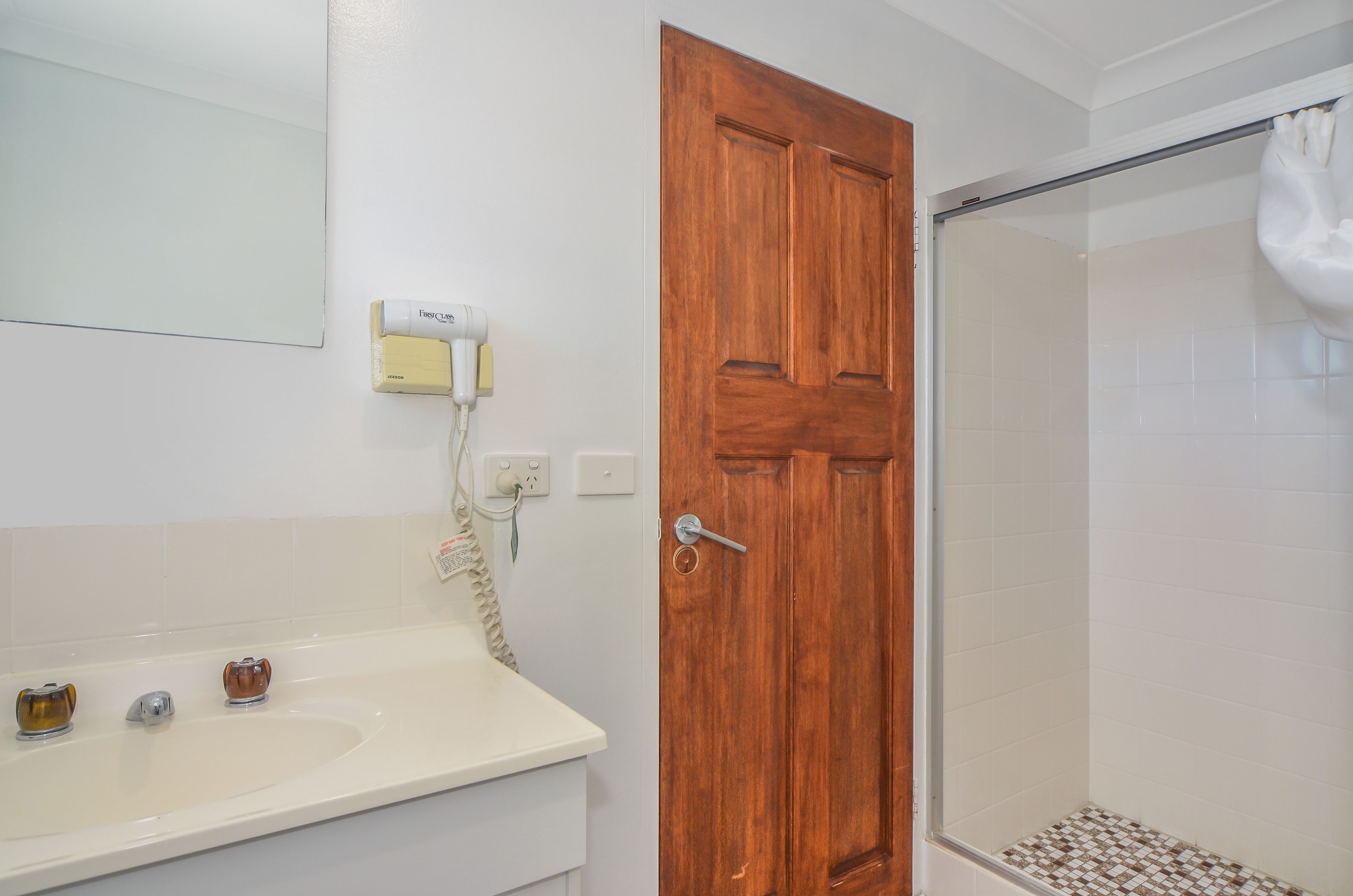 Un baño con puerta de madera, un lavabo blanco con dos grifos y una zona de ducha con puerta de cristal.