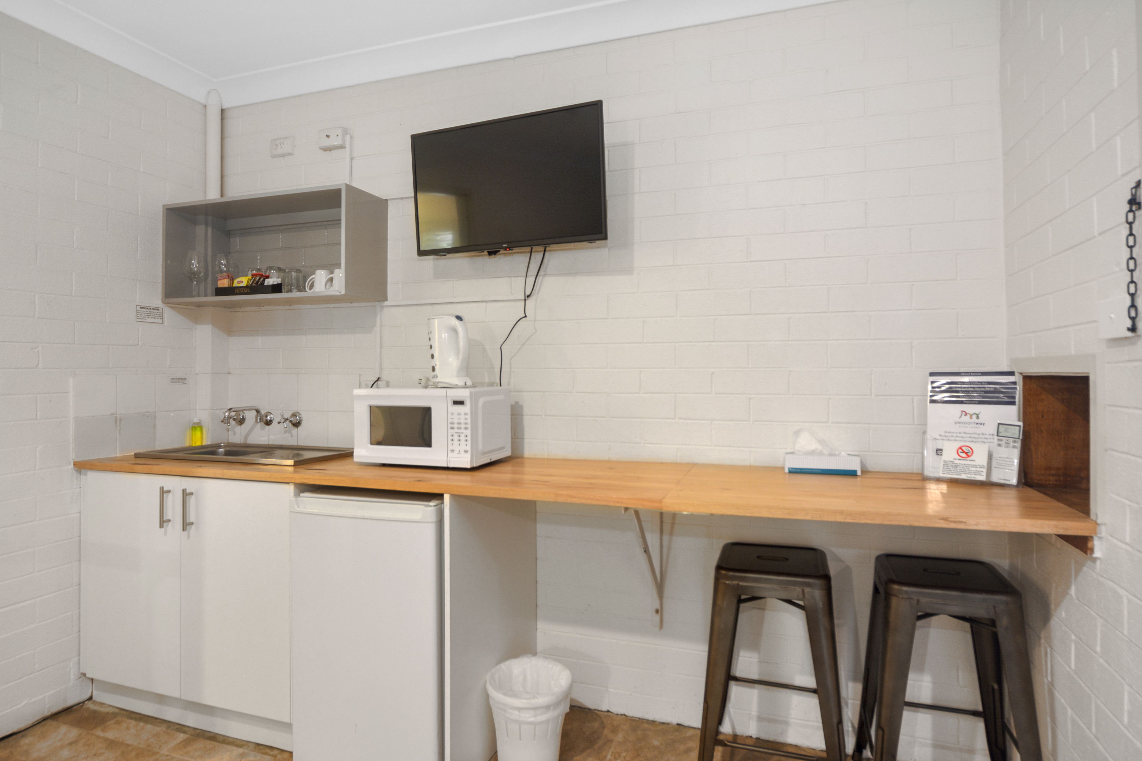 Una cocina compacta con armarios blancos, microondas, fregadero, refrigerador pequeño, encimera de madera, televisor montado en la pared y dos taburetes.