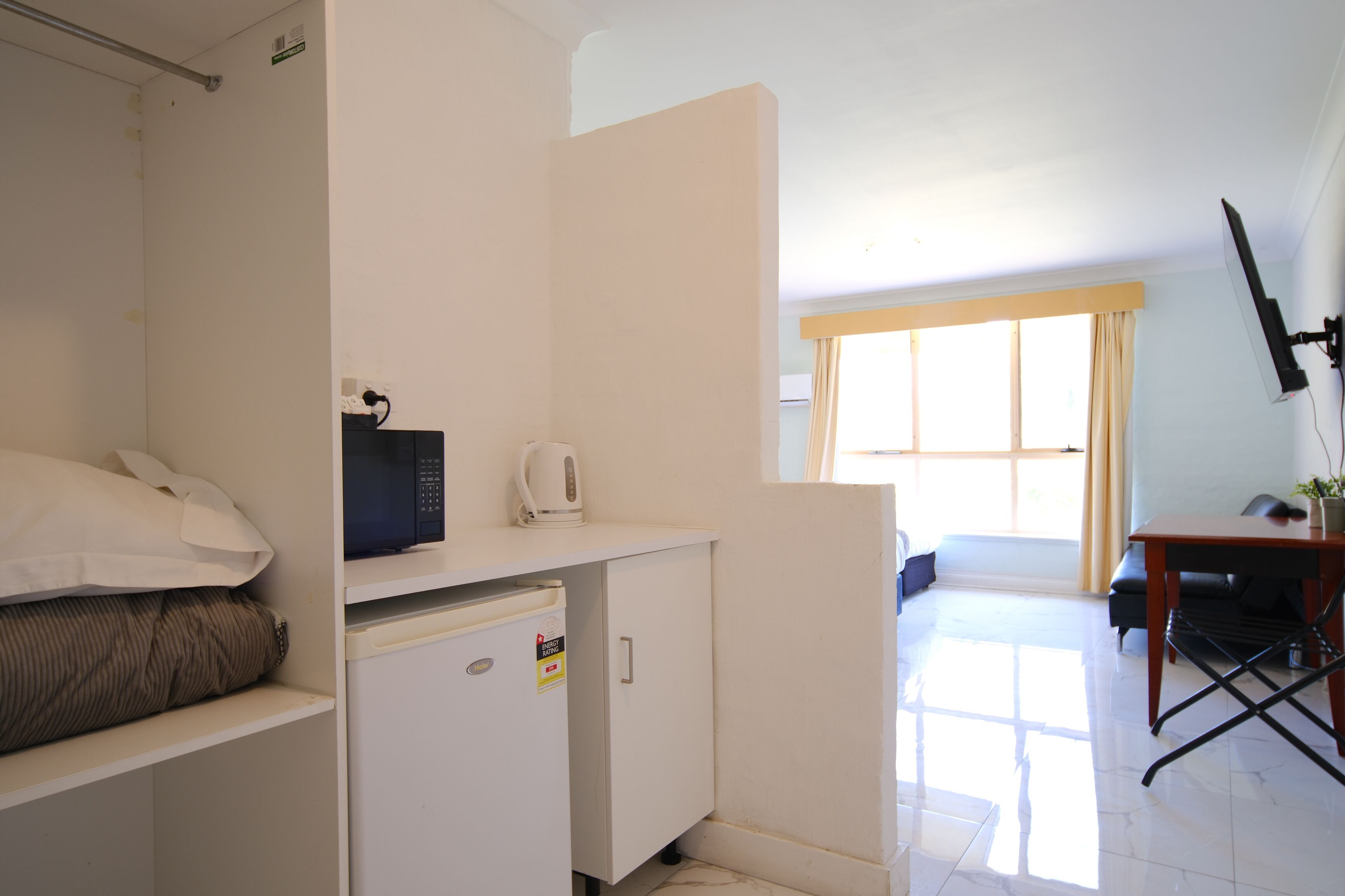 A compact hotel room with a kitchenette, a bed, a small desk, and a window with curtains.