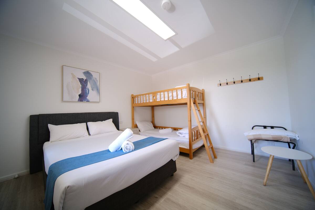 A bedroom with a bunk bed, a single bed, a bedside table, a wall-mounted coat rack, and a ceiling light.