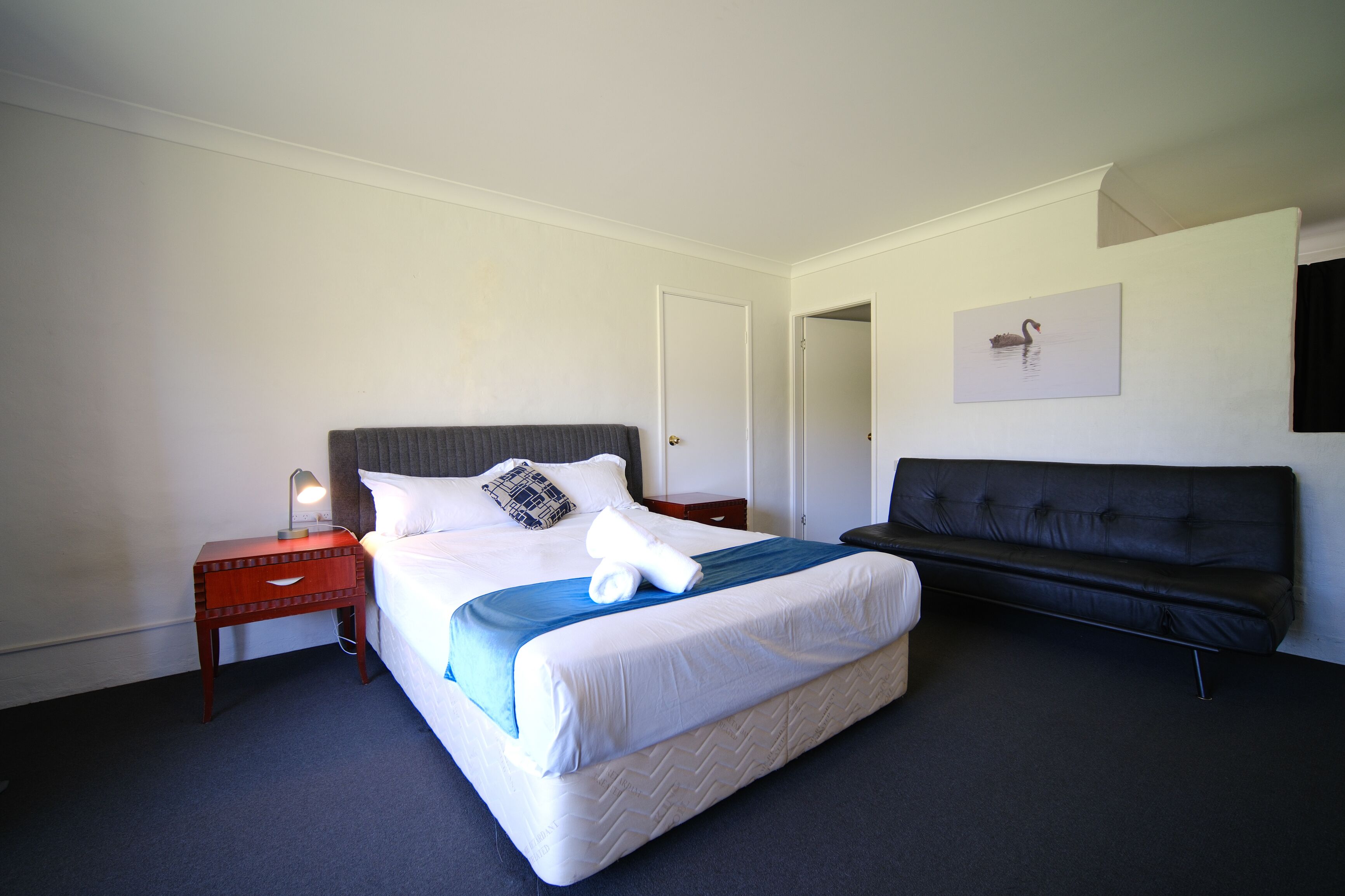 Una habitación de hotel con cama, una mesita de noche con lámpara, un sofá negro y un cuadro colgado en la pared.