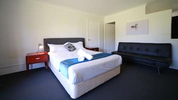 Una habitación de hotel con cama, una mesita de noche con lámpara, un sofá negro y un cuadro colgado en la pared.