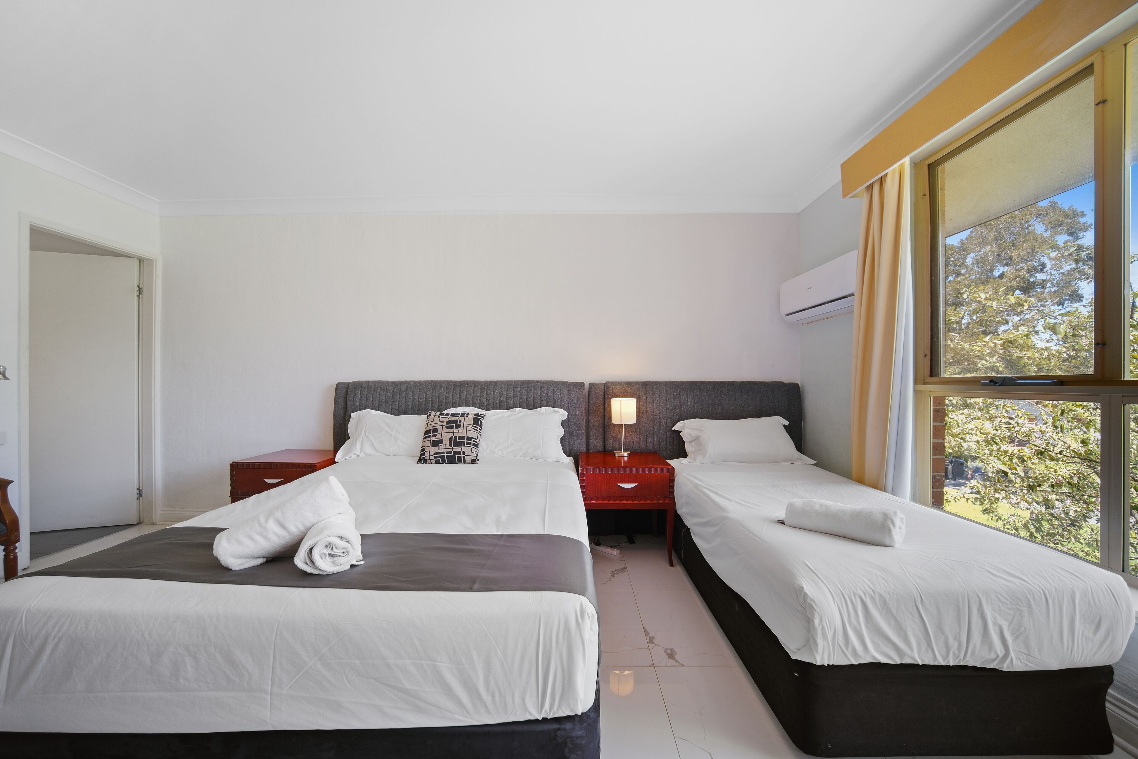 Habitación de hotel con dos camas, una mesita de noche, una lámpara y una ventana con vistas a árboles.