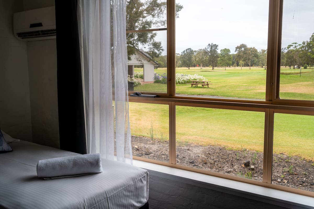 A room with a large window, a bed with a pillow, and a view of a grassy area with trees and a building.