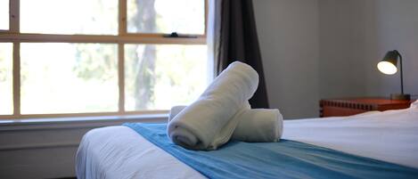 A neatly made bed with white linens and a blue bedspread, a window with curtains, and a bedside lamp.