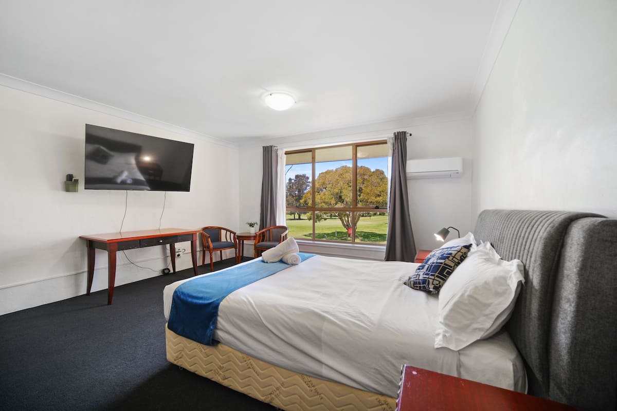 A hotel room with a bed, a desk, a chair, a TV, and a window with a view of trees.