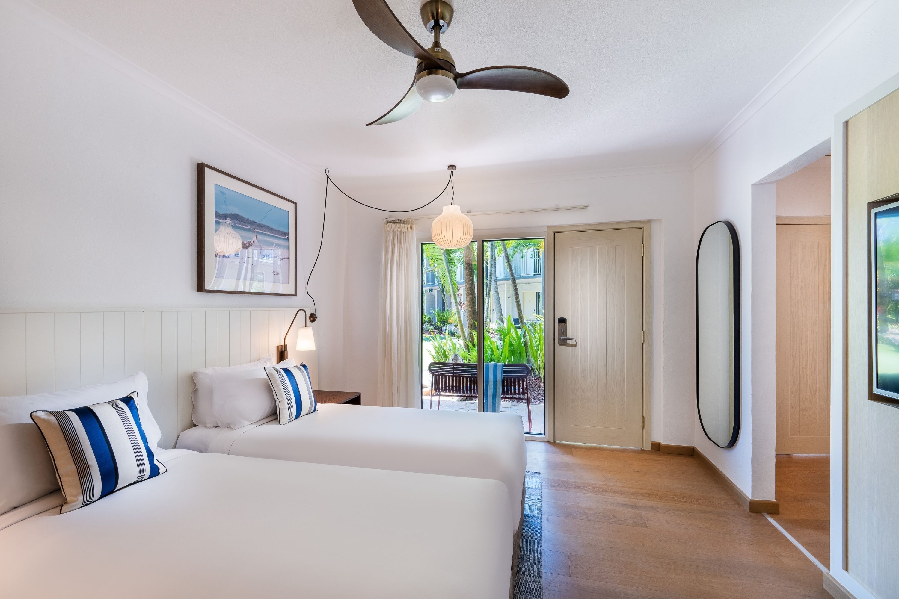 Hotelkamer met twee bedden, een plafondventilator, een groot raam met uitzicht op groen en een deur naar een buitenruimte.