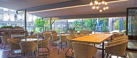 An indoor dining area with wicker chairs and tables, large windows, and a view of greenery outside.
