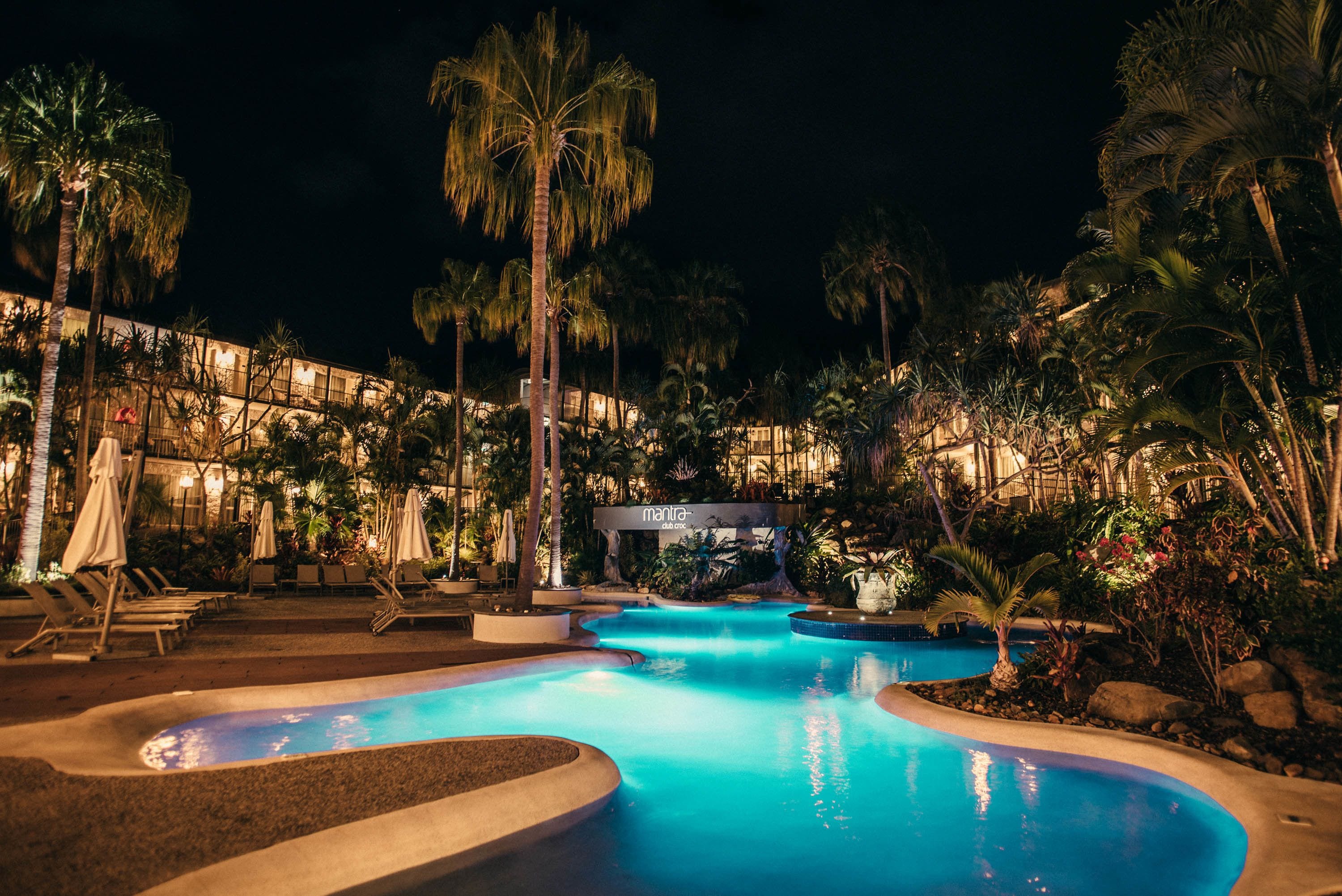 Zona de piscina iluminada de noche con sillones de descanso y palmeras.