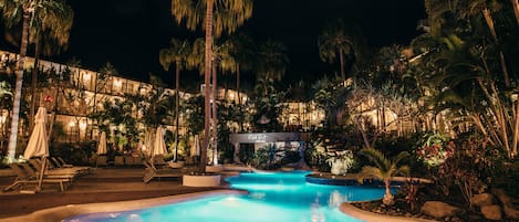 Zona de piscina iluminada de noche con sillones de descanso y palmeras.
