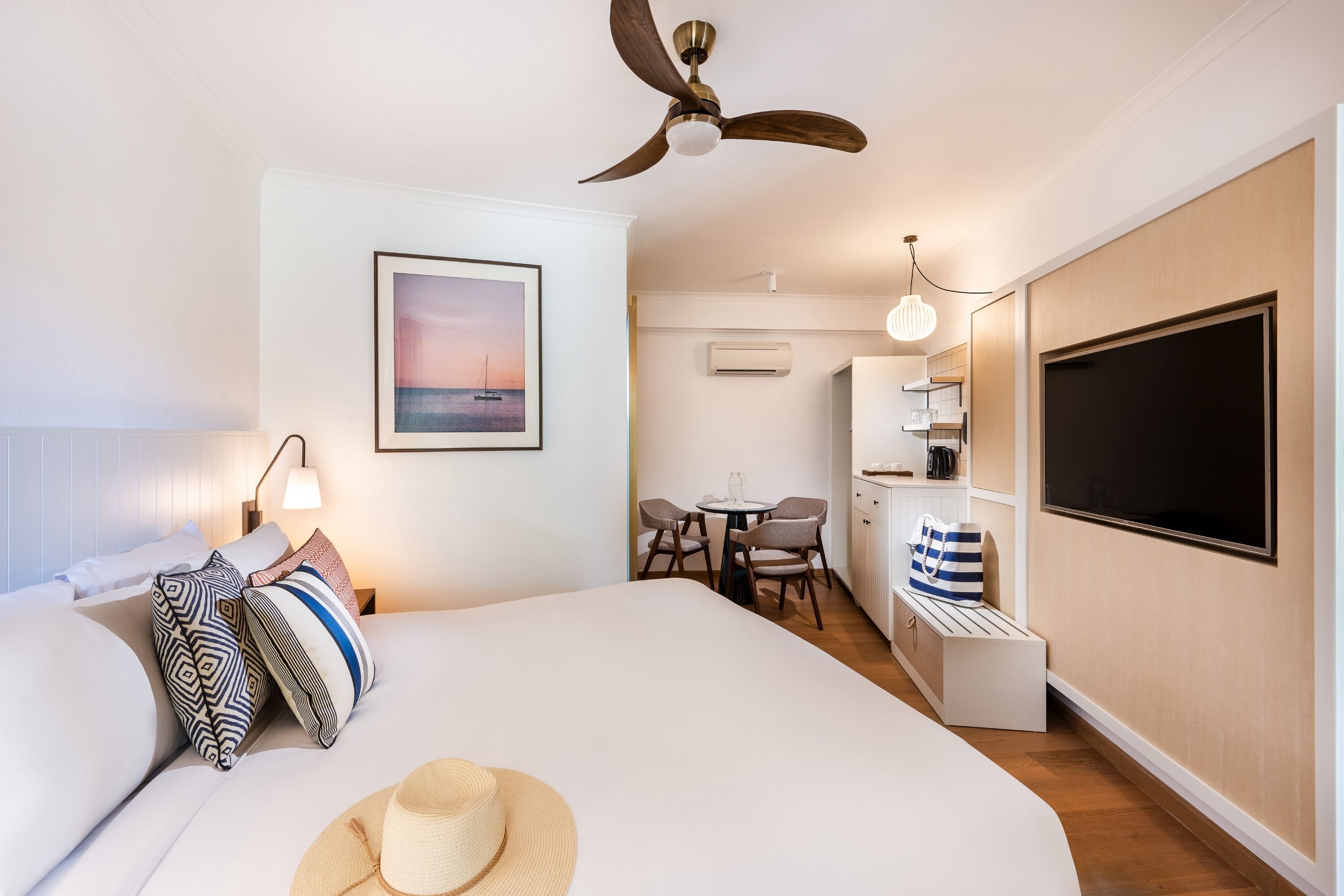 A modern hotel room with a large bed, a ceiling fan, a dining area with a table and chairs, and a TV mounted on the wall.