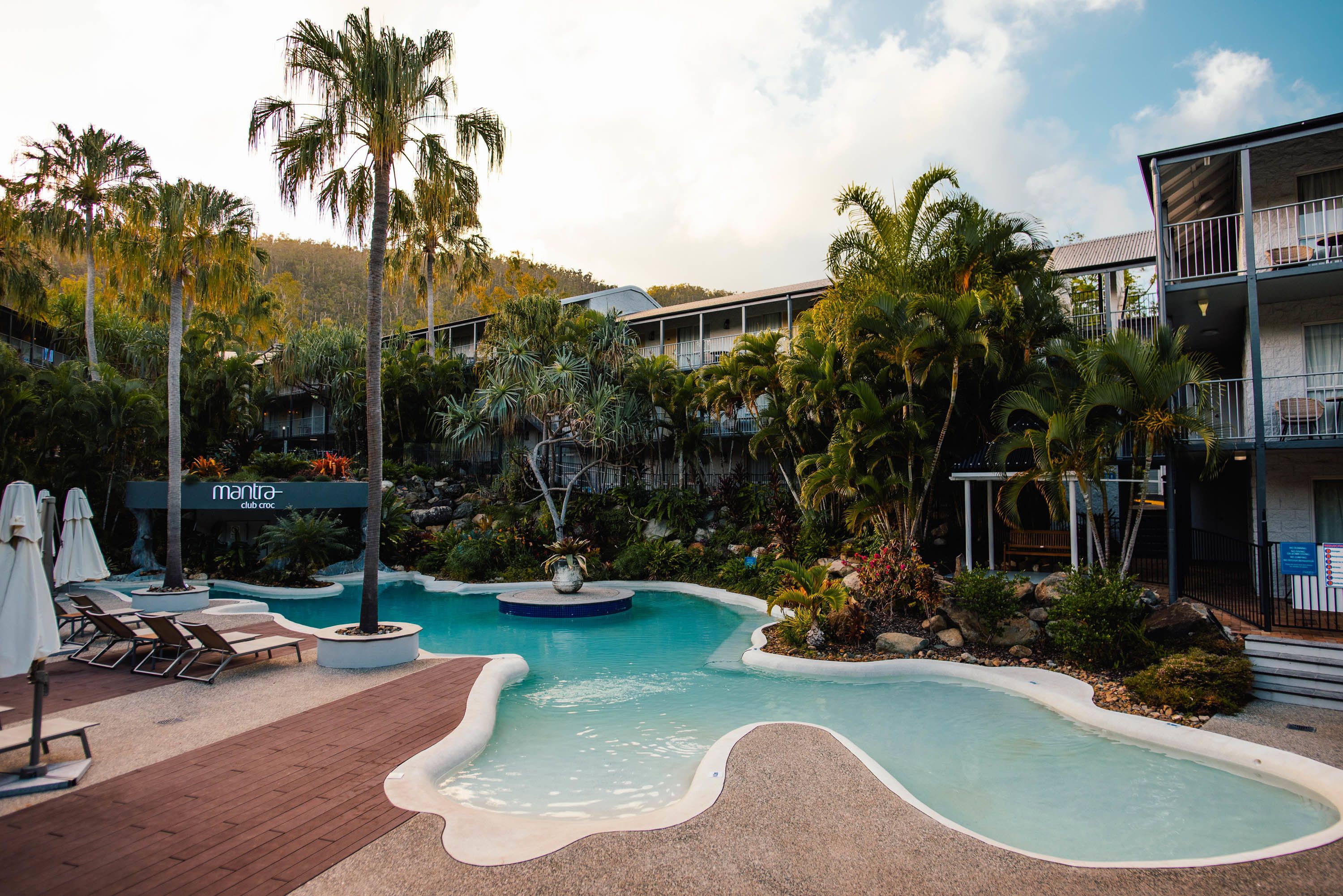 Zwembadgedeelte van een resort met ligbedden, palmbomen en een bordje met 'Mantra Club Croc' erop.