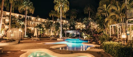 Zona de piscina en un resort por la noche, con varias piscinas, palmeras y sillas de descanso.