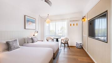 Habitación de hotel con dos camas, ventilador de techo, ventana con cortinas, una mesa con sillas y televisor montado en la pared.