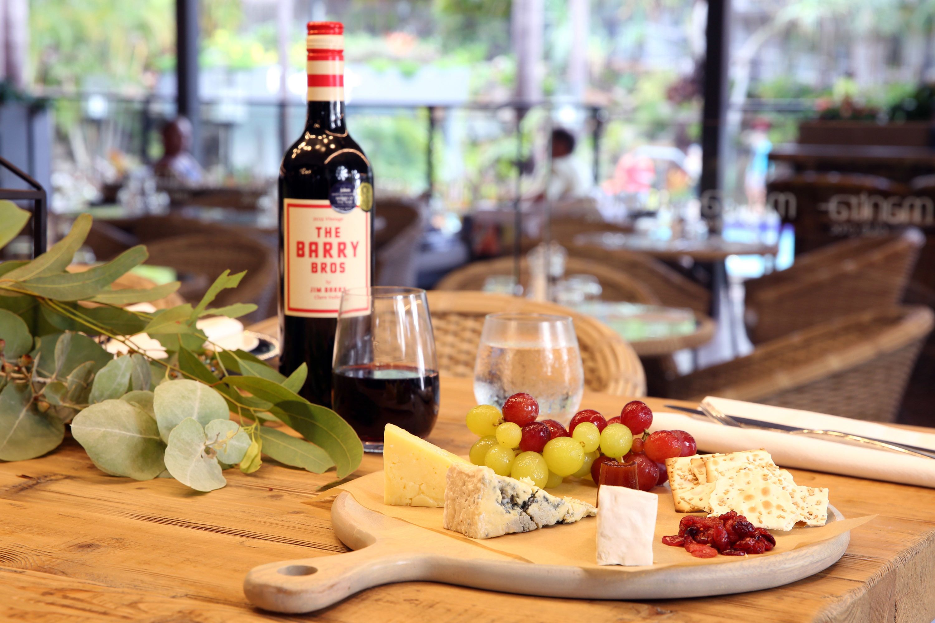 A wooden table with a variety of cheeses, grapes, and a bottle of wine, set in a restaurant with a view of the outdoors.