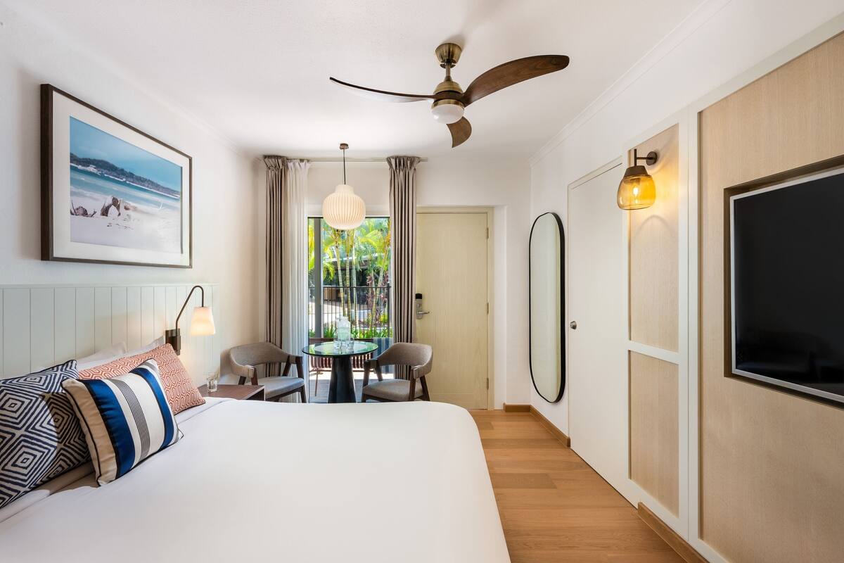 A hotel room with a bed, a ceiling fan, a TV, a dining table with chairs, and a view of the outdoors.