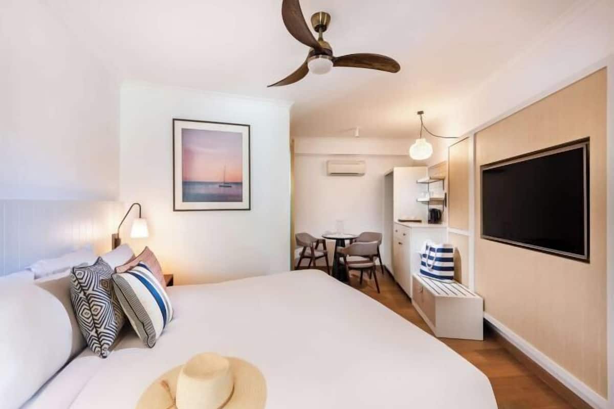 A hotel room with a bed, a ceiling fan, a desk with a chair, a TV, and a picture on the wall.