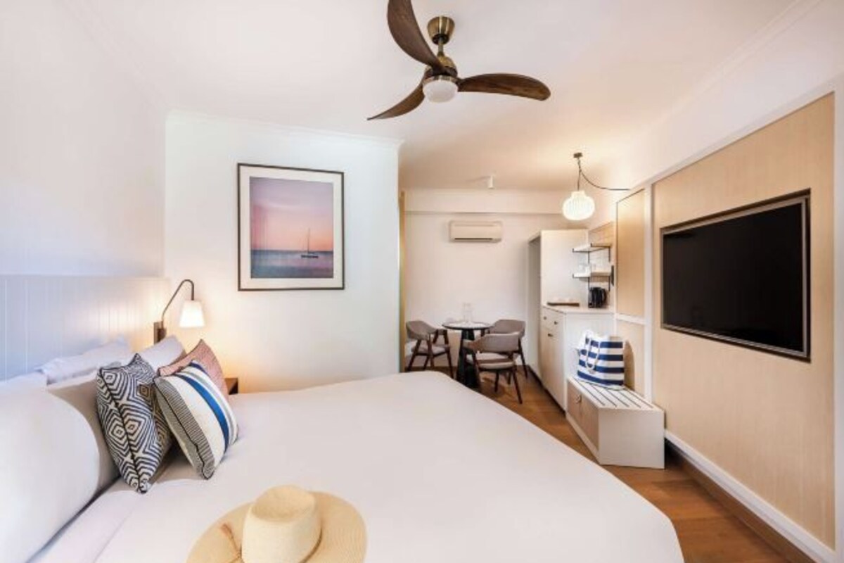 A hotel room with a bed, a ceiling fan, a desk with a chair, a TV, and a picture on the wall.
