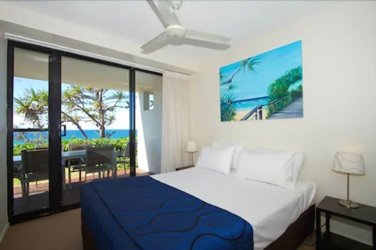 A hotel room with a bed, a ceiling fan, a painting of a beach, and a view of the ocean through a sliding door.