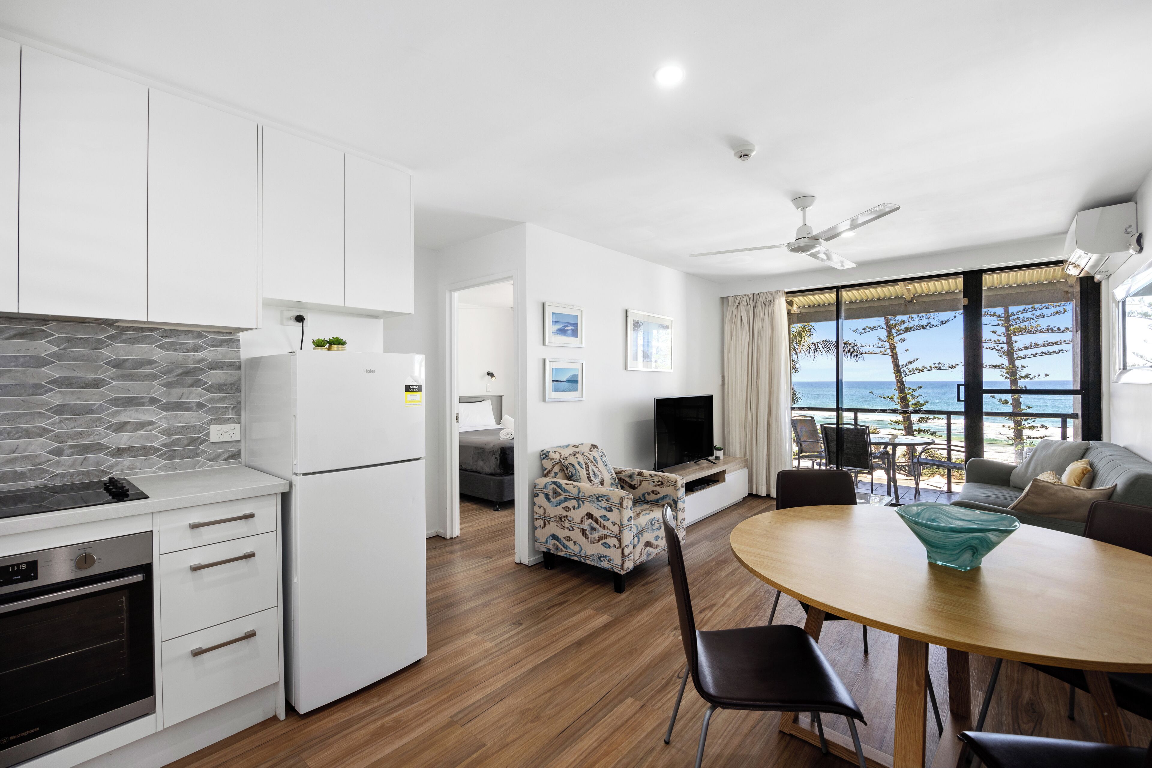 A modern kitchen with a built-in oven, a dining area with a round table, and a view of the ocean through large windows.