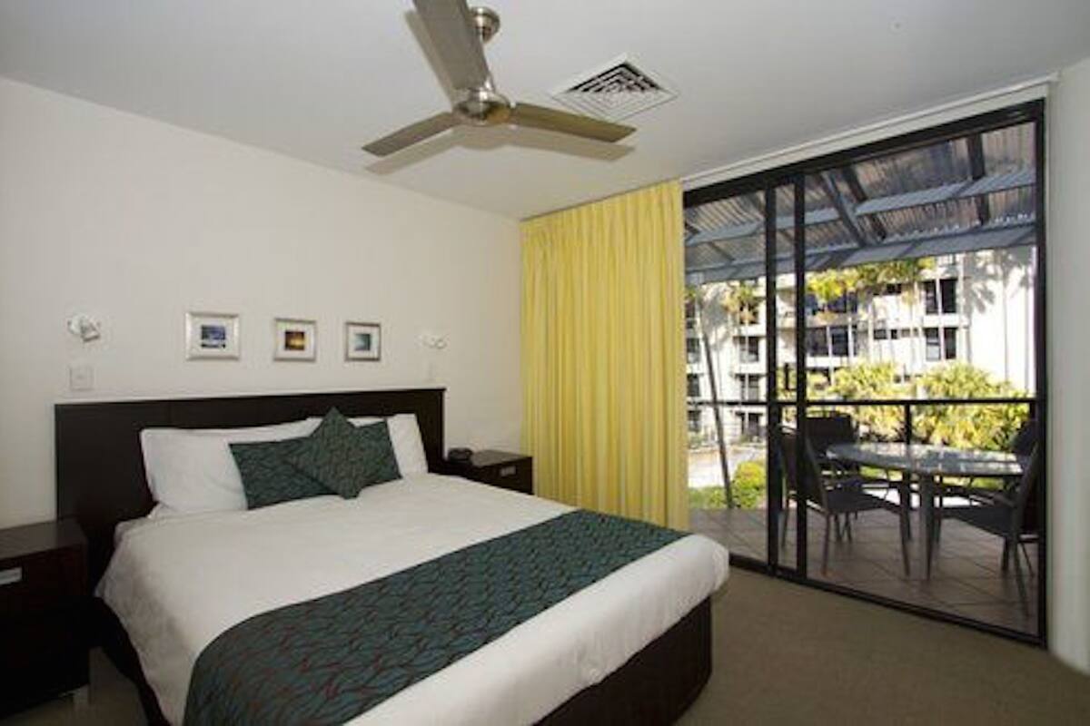 A hotel room with a bed, a ceiling fan, a window with yellow curtains, and a balcony with outdoor furniture.