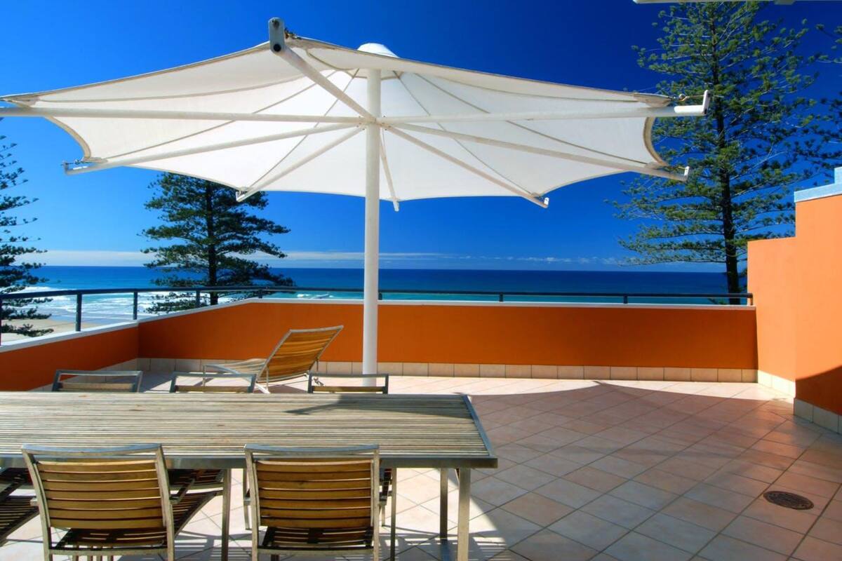 A terrace with a table and chairs, a large umbrella, and a view of the sea.
