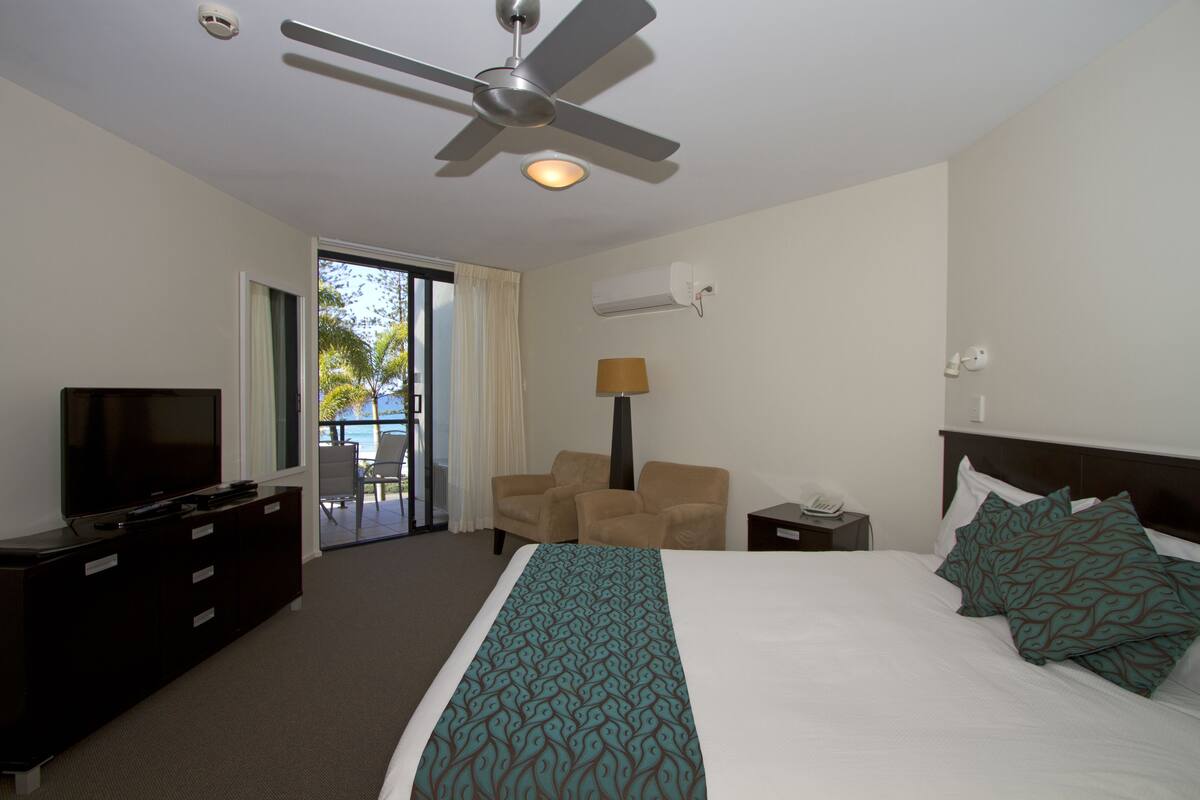 A hotel room with a bed, a TV, a ceiling fan, a desk with a lamp, and a chair.