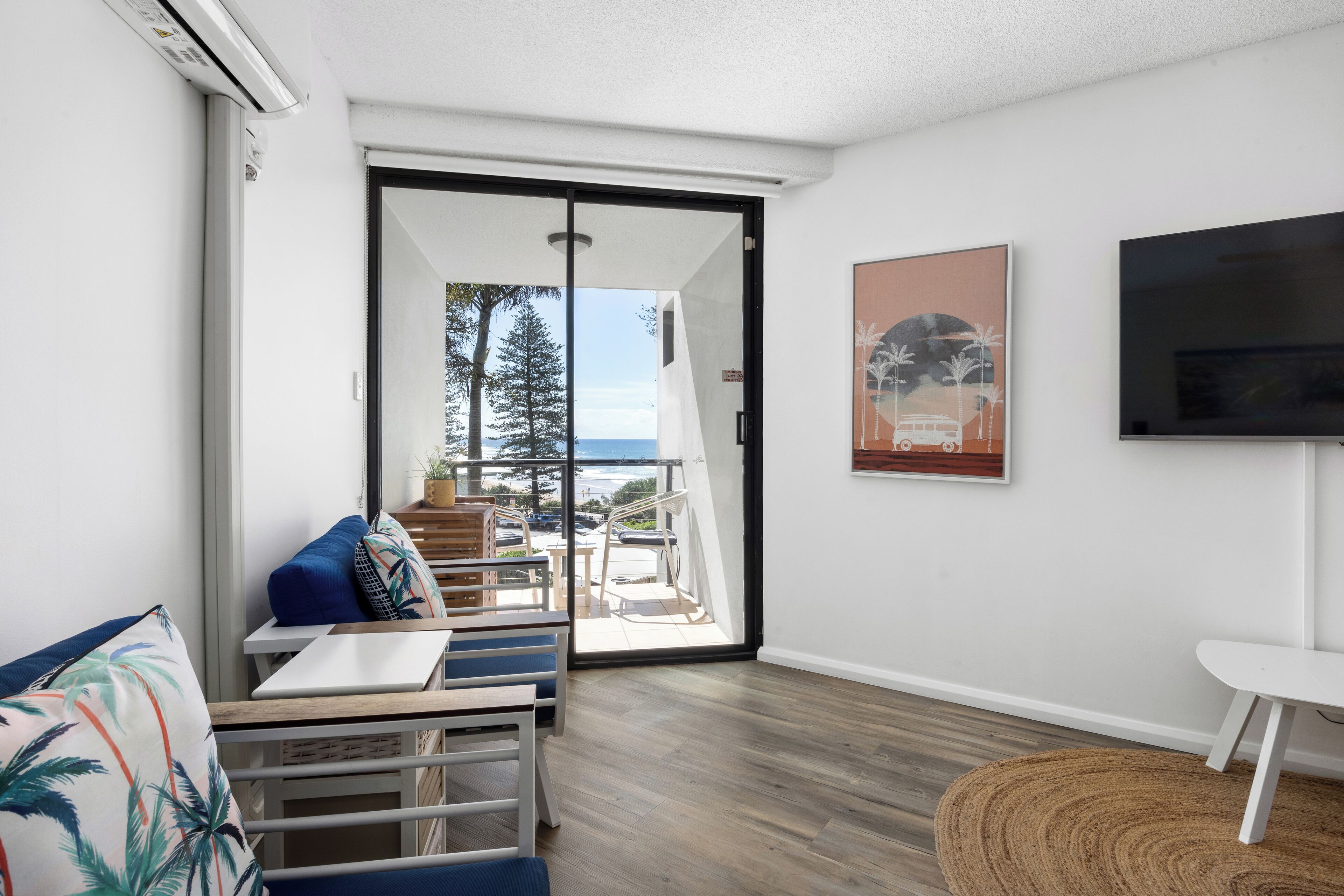 Ein modernes Wohnzimmer mit einer Couch, einem Couchtisch und einem Fernseher. Eine Glastür führt auf einen Balkon mit Blick auf das Meer und Bäume.