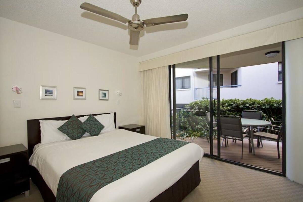 A hotel room with a bed, a ceiling fan, a nightstand, a window with curtains, and a view of an outdoor patio with a table and chairs.