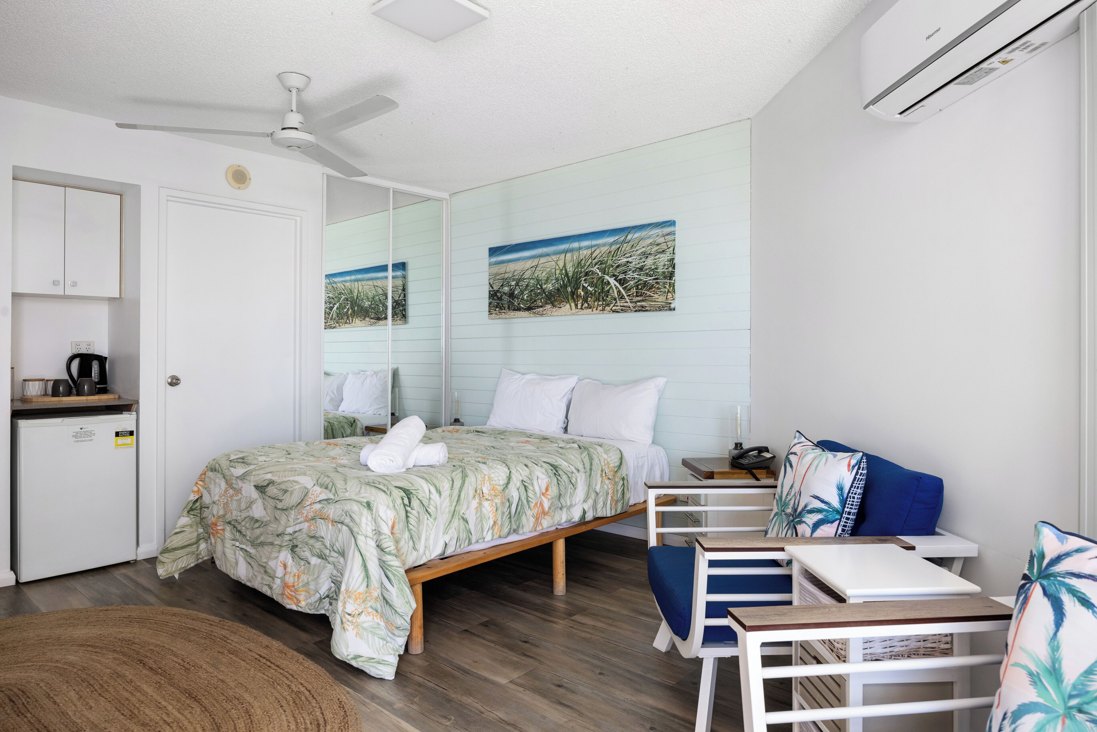 Un dormitorio con cama, escritorio, silla, ventilador de techo y una pequeña kitchenette.