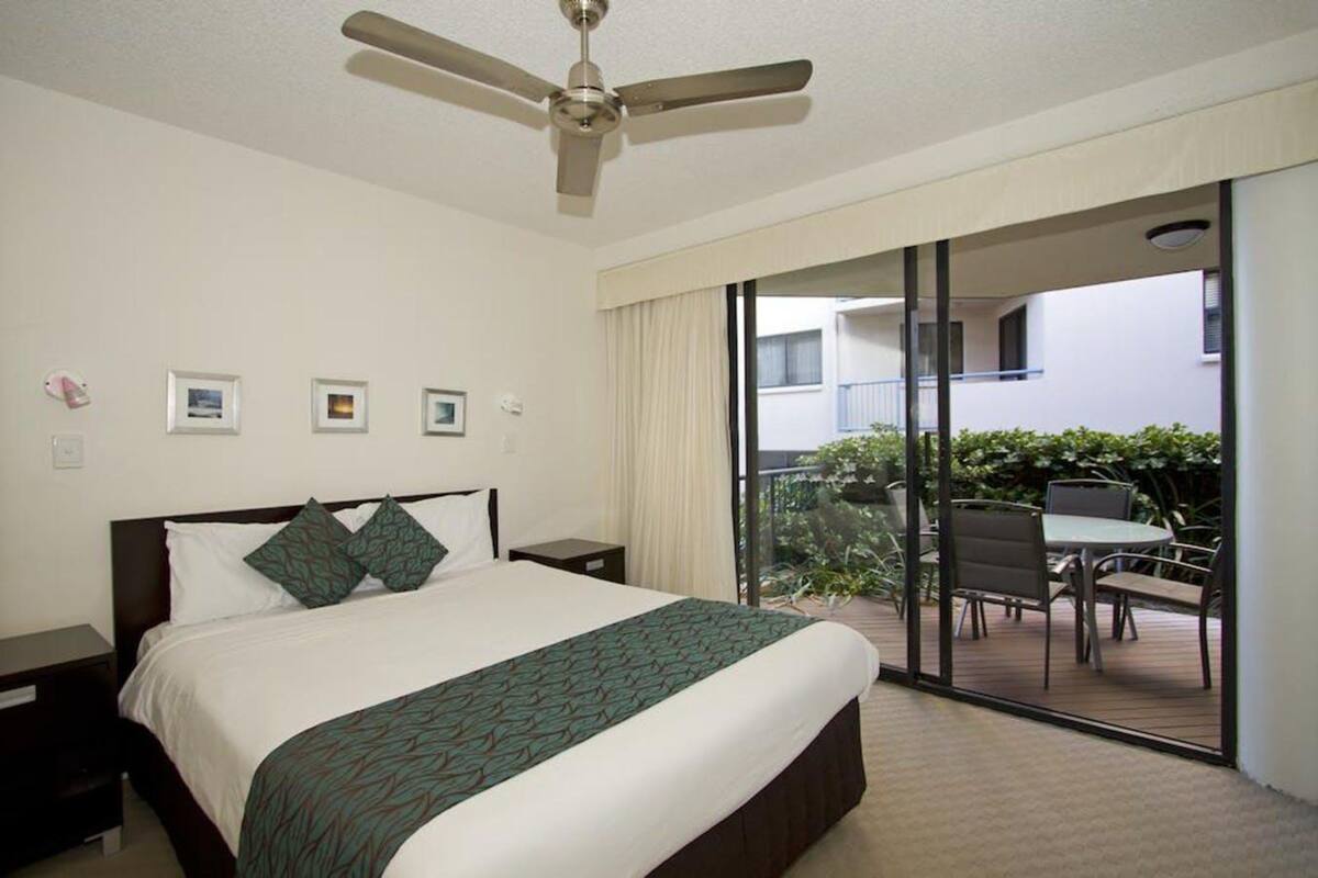 A hotel room with a bed, a ceiling fan, a balcony with a table and chairs, and a view of the building outside.