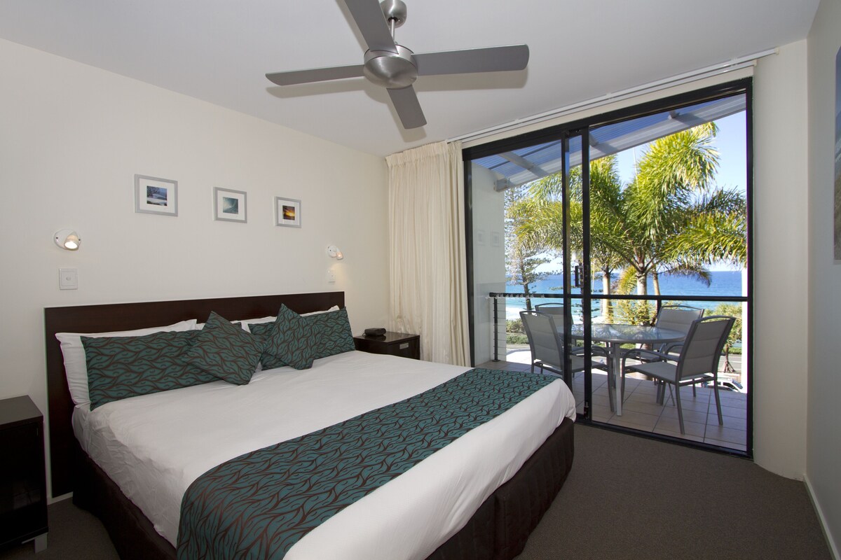 A hotel room with a bed, a ceiling fan, a balcony with a table and chairs, and a view of the sea.