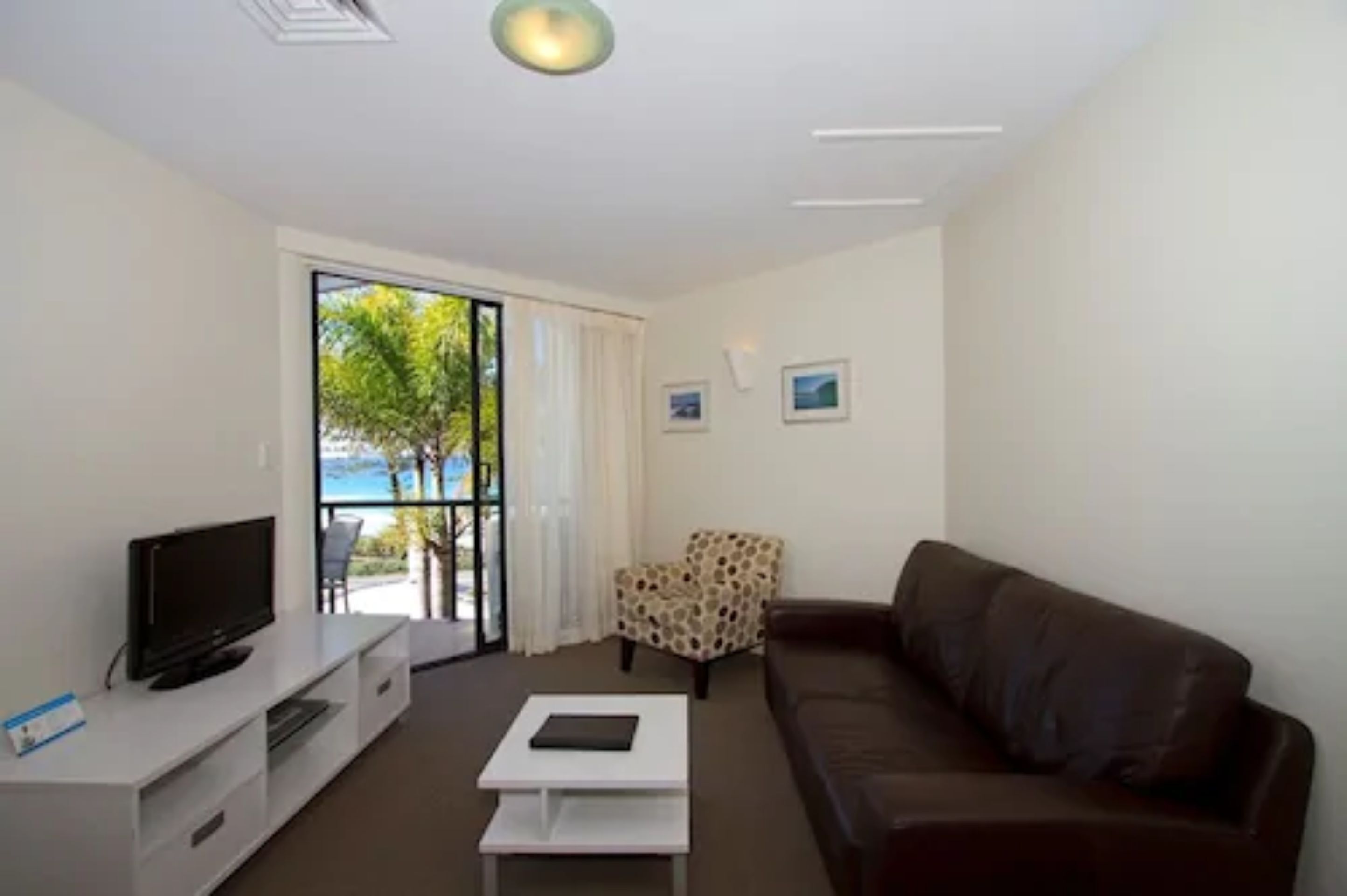 A modern living room with a flat-screen TV, a white coffee table, a brown leather sofa, and a patterned armchair.