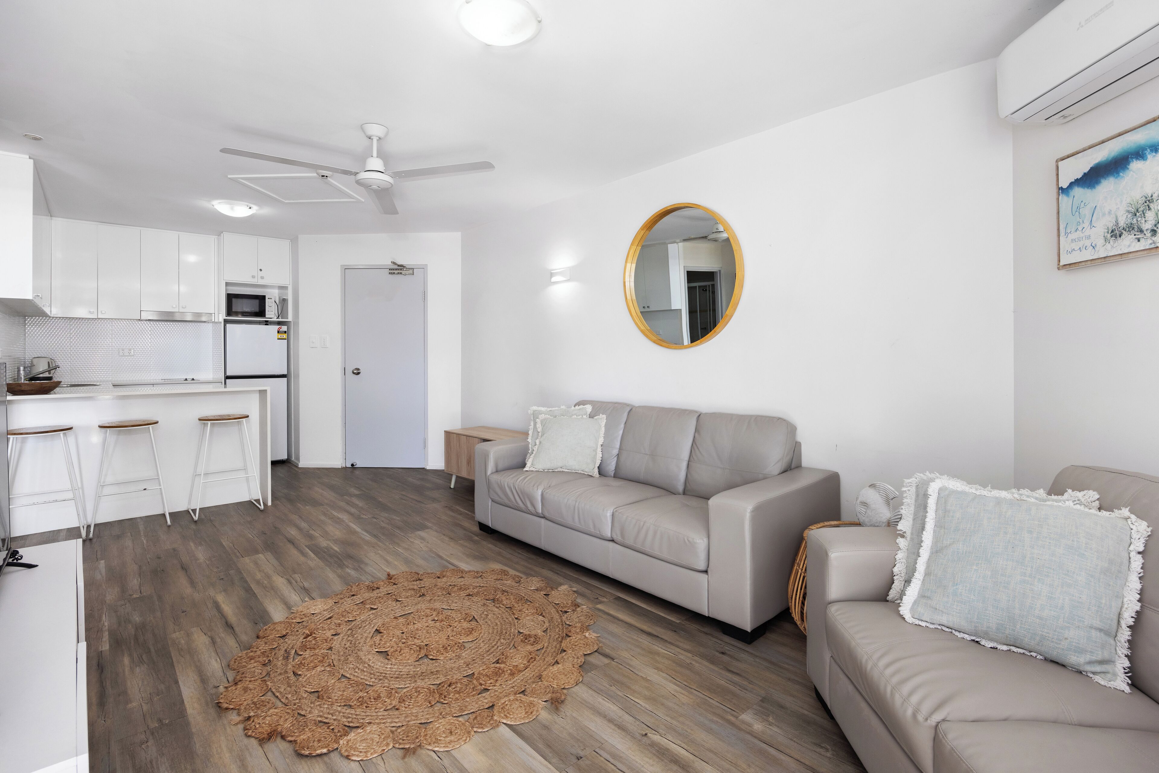 A modern living room with a grey sofa, a round mirror, and a kitchen area with white cabinets and a microwave.