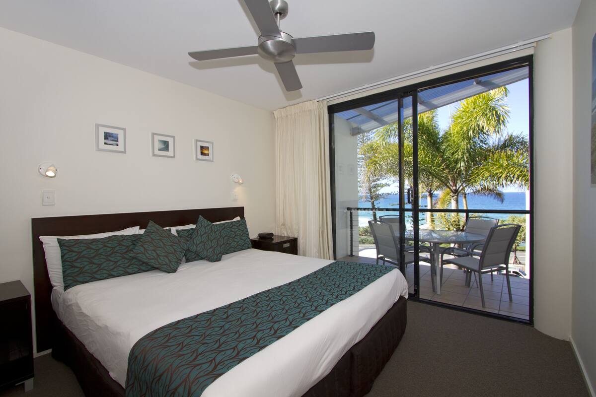 A hotel room with a large bed, a ceiling fan, a balcony with a table and chairs, and a view of the ocean.