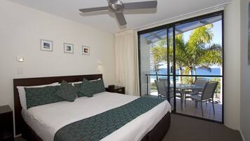A hotel room with a large bed, a ceiling fan, a balcony with a table and chairs, and a view of the ocean.