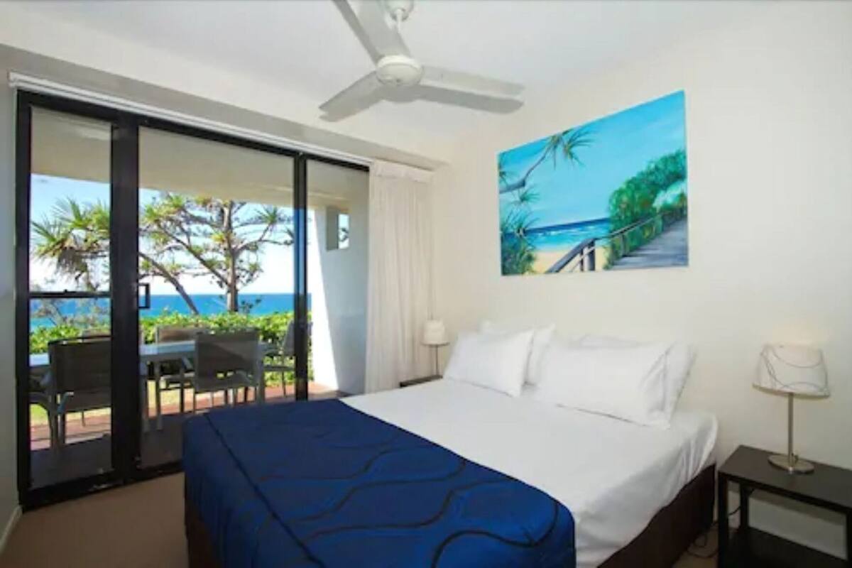 A hotel room with a bed, a painting of a beach, a ceiling fan, and a view of the ocean through a sliding door.