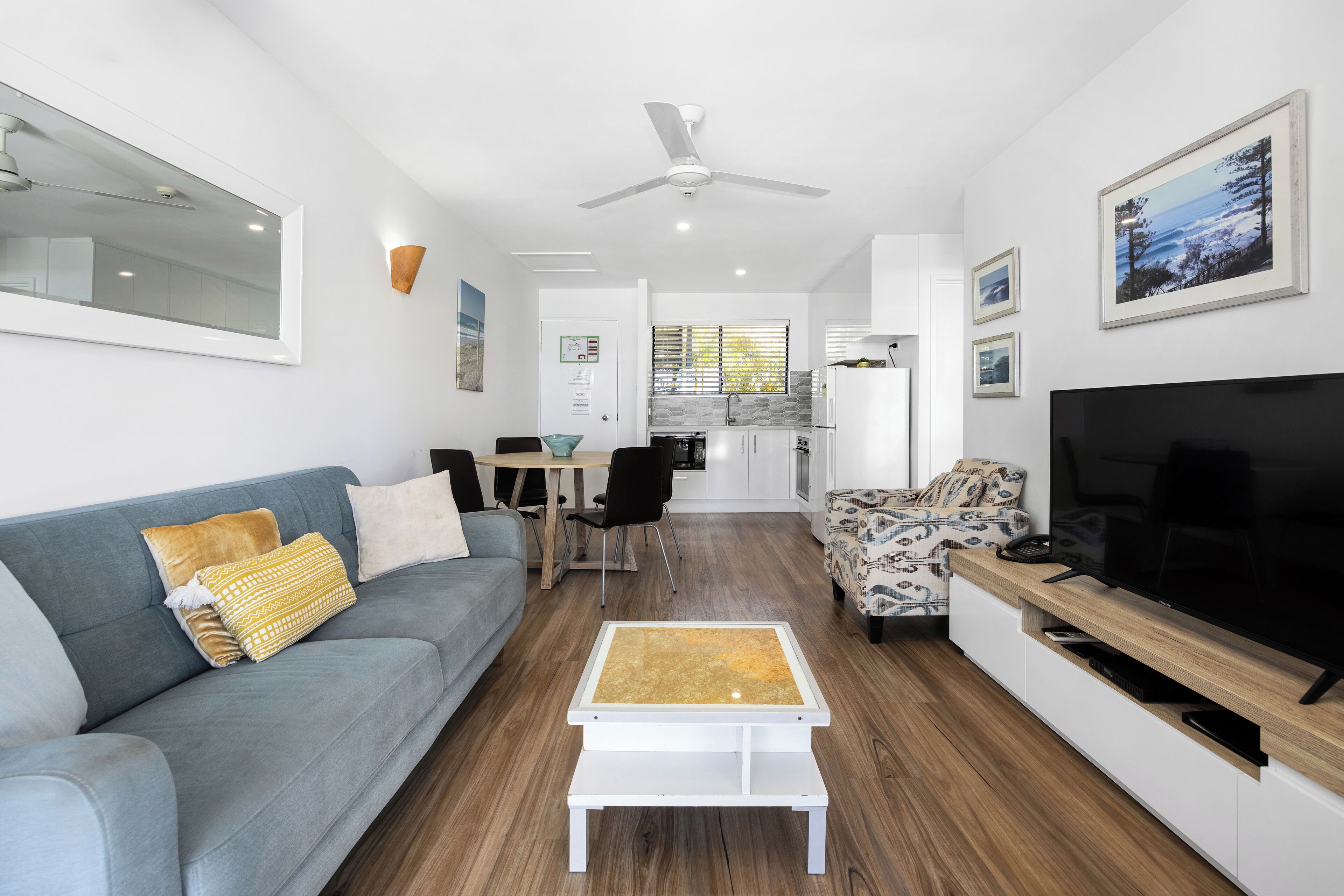 A modern living room with a grey sofa, a coffee table, a TV, and a kitchen in the background.
