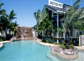A hotel with a pool, palm trees, and a waterfall feature. - Runaway Bay Motor Inn (Runaway Bay)