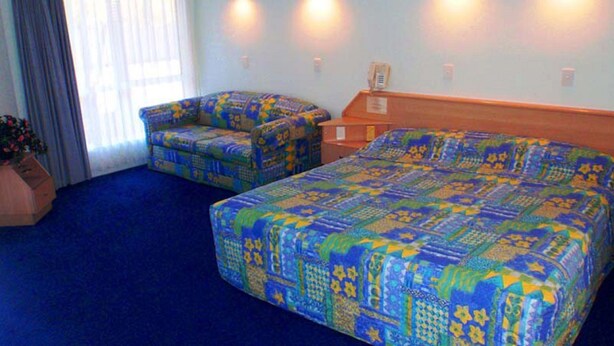 A hotel room with a large bed, two armchairs, a wooden nightstand, and a potted plant. - Runaway Bay Motor Inn (Runaway Bay)