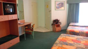 Ein Hotelzimmer mit Schreibtisch, Sessel, Fernseher, einem Bett mit bunter Decke, einer kleinen Holzkabine mit Blumen und einer Tür.