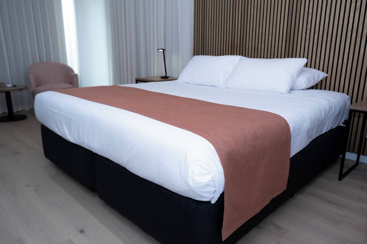Quarto de hotel moderno com uma cama grande, uma poltrona, uma mesinha de cabeceira com abajur e painel de parede de madeira.
